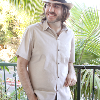 Man in handmade sandy colored short sleeved button up shirt leaning against a railing and wearing a straw fedora