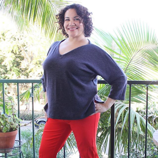 Curly haired brunette in a handmade grey v-neck sweater and red pants standing against a green railing