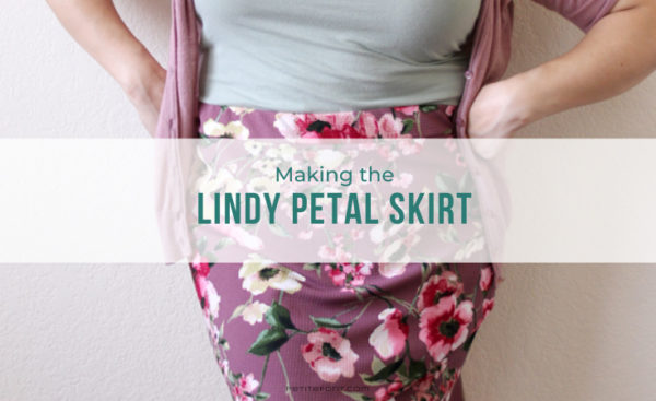 Close up image of a woman leaning against a wall with her hands on her hips, wearing a light green tank top, pink cardigan, and mauve floral skirt. Teal text overlay reads Making the Lindy Petal Skirt, Petite Font dot com