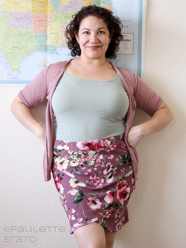 Image of a curly-haired Latina woman leaning against a wall with a US flag poster on it, her hands on her hips, wearing a light green tank top, pink cardigan, and mauve floral skirt
