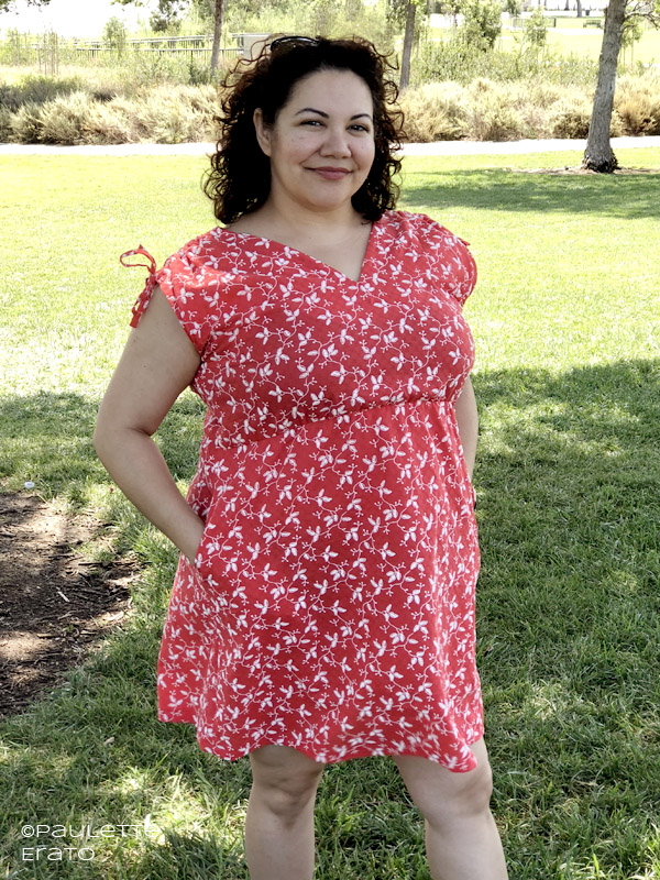 Curly haired brunette in a handmade red cotton Seamwork Patterns Kimmy dress. The fabric has faux embroidery and she is standing in the shade of a large grassy field.