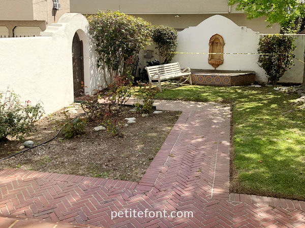 Garden area with brick walkway between foliage and an empty bench next to a dry fountain in Alamitos Beach, Long Beach