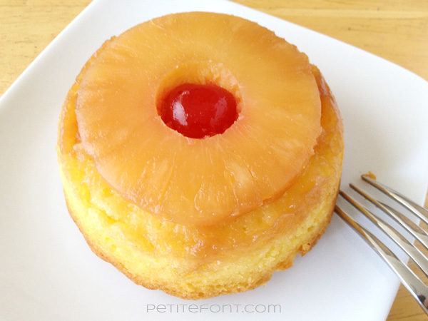 Single round serving of pineapple upside down cake on a white plate, topped with a cherry and a fork to the side