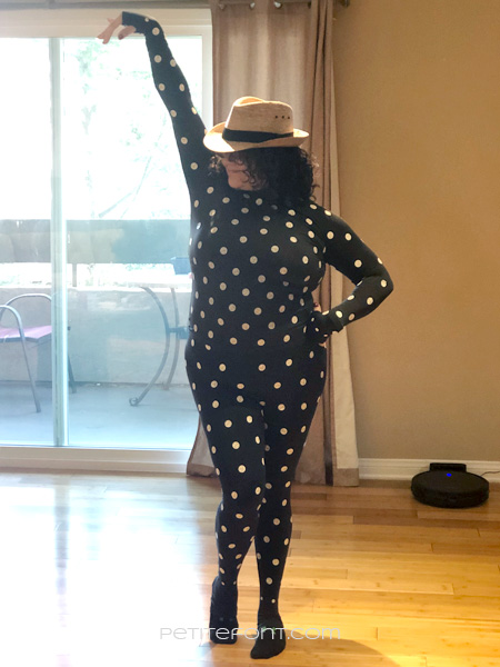 Front view of woman in Zozo suit, a black stretchy body suit covered in white dots, with a straw hat and one arm up in the air