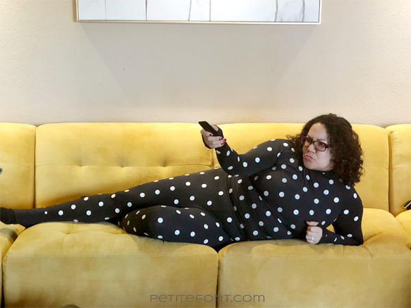 Woman stretched out on a yellow couch holding a remote and scowling at something off to the left, while wearing the Zozo suit (a black stretchy body suit covered in white polka dots)