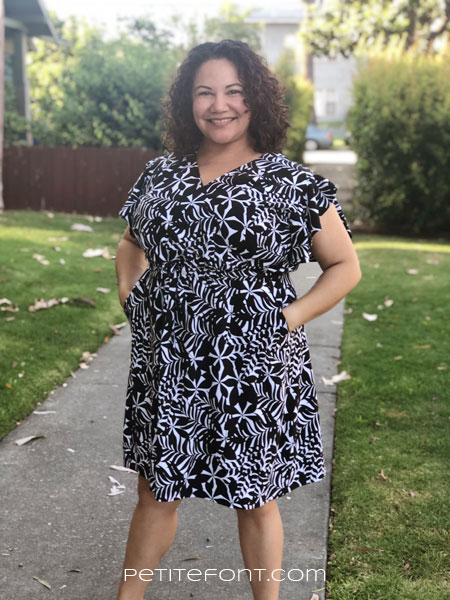 Front view of a curly haired brunette with her hands in the pockets of a shortened and narrowed Orchid dress in black and white floral fabric