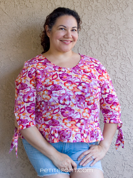 Curly haired brunette leaning against a stucco wall wearing a handmade Franki top from Wearable Studio patterns, in floral cotton gauze