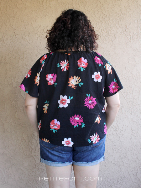 Back view of a curly haired brunette leaning against a stucco wall wearing a handmade Seamwork Loretta in black floral cotton gauze and jean shorts. Website address is petite font dot com.