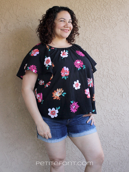 Curly haired brunette leaning against a stucco wall wearing a handmade Seamwork Loretta in black floral cotton gauze and jean shorts. Website address is petite font dot com.