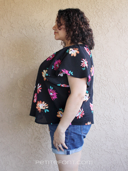 Side view of a curly haired brunette leaning against a stucco wall wearing a handmade Seamwork Loretta in black floral cotton gauze and jean shorts. Website address is petite font dot com.