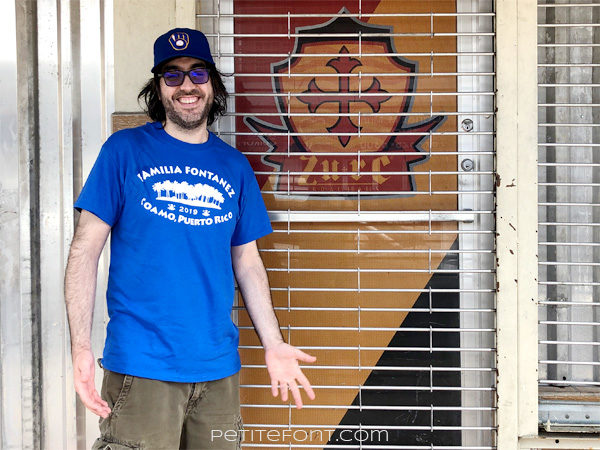 Bearded man in blue shirt and baseball hat standing in front of a closed breweryi