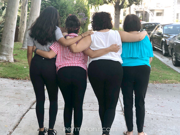 Back view of 4 women wearing black leggings as pants without their shirts covering their butts, with their arms around each other