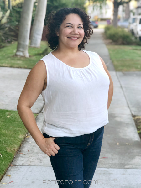 Latina curly haired woman in a white sleeveless hacked Daisy blouse and skinny jeans facing forward with one hand behind her and another with her thumb hooked into her front pocked