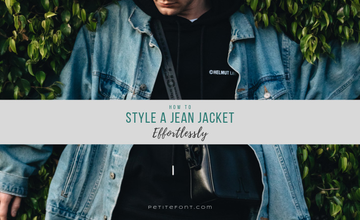 A person in black wearing a light blue vintage denim jacket leaning against bushes looking down with text overlay that reads "how to style a jean jacket effortlessly" petite font dot com