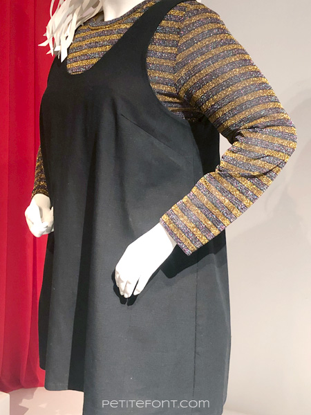 Cropped image of a mannequin wearing a baggy black pinafore dress over a striped sweater, both of which are far too large for the mannequin, from FIDM's 13th Art of Television Costume Design exhibit