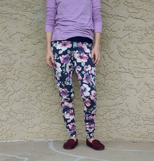 Cut off image of a person in lavender long sleeve shirt, floral Rad Patterns jammers, and dark moccasins leaning against a beige stucco wall