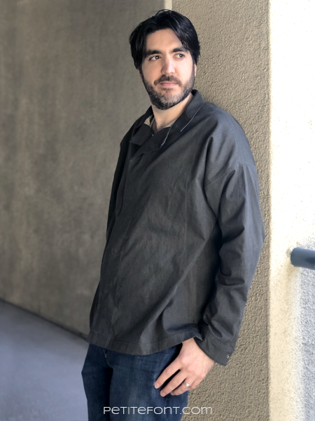Dark haired man wearing a dark charcoal grey Ilford jacket as a shirt, with jeans, leaning up against a beige stucco wall