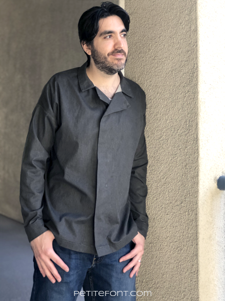Dark haired man wearing a dark charcoal grey Ilford jacket as a shirt, with jeans, leaning up against a beige stucco wall