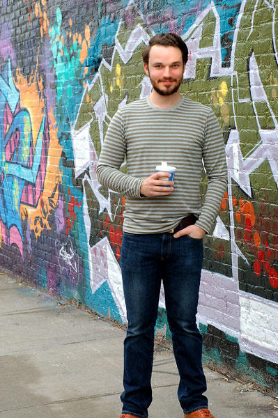 Man in a striped long sleeve shirt and black pants holding a coffee cup and standing in front of a mural, modeling the Liesl & Co Metro t-shirt