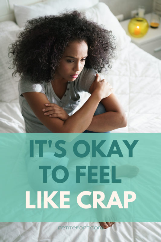 Black woman sitting on her bed looking upset with text overlay that reads It's Okay to feel Like Crap PetiteFont.com