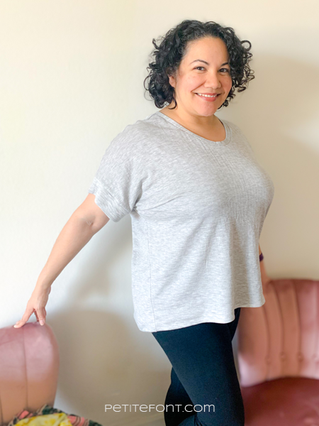 Curly haired Latina in a light grey handmade Lou Box Top and black leggings standing in front of two pink chairs