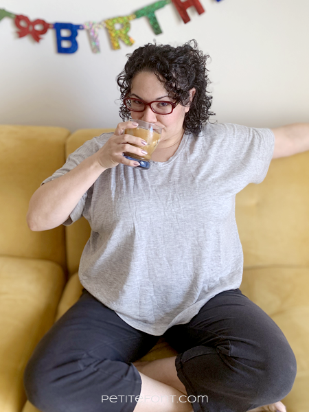 Paulette in a hacked grey Lou Box Top sitting on the couch under a Happy Birthday banner sipping an iced coffee