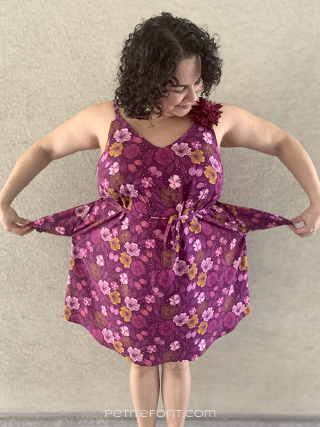 Curly haired latina woman in a floral Sangria Misty Cami dress showing off the pockets. Text overlay reads PetiteFont.com