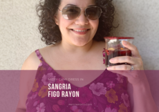 Close up of a curly haired Latina woman in sunglasses and a floral purple dress holding a glass filled with sangria. Text overlay in a purple box reads "Misty Cami Dress in Sangria Figo Rayon, PetiteFont.com"