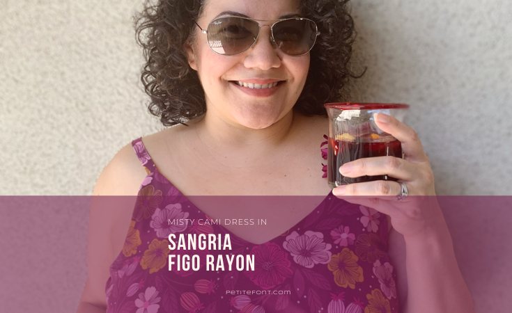 Close up of a curly haired Latina woman in sunglasses and a floral purple dress holding a glass filled with sangria. Text overlay in a purple box reads "Misty Cami Dress in Sangria Figo Rayon, PetiteFont.com"