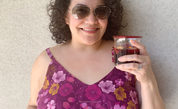 Close up of a curly haired Latina woman in sunglasses and a floral Sangria Misty Cami dress holding a glass filled with sangria. Text overlay reads PetiteFont.com
