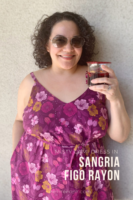 Curly haired latina woman in sunglasses and a purple floral dress holding a glass filled with sangria. Text overlay reads "Misty Cami Dress in Sangria Figo Rayon, PetiteFont.com"