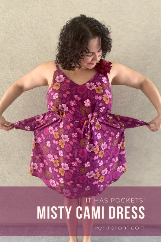 Curly haired latina woman in a floral Sangria Misty Cami dress showing off the pockets. Text overlay "It has pockets! Misty Cami Dress reads PetiteFont.com"