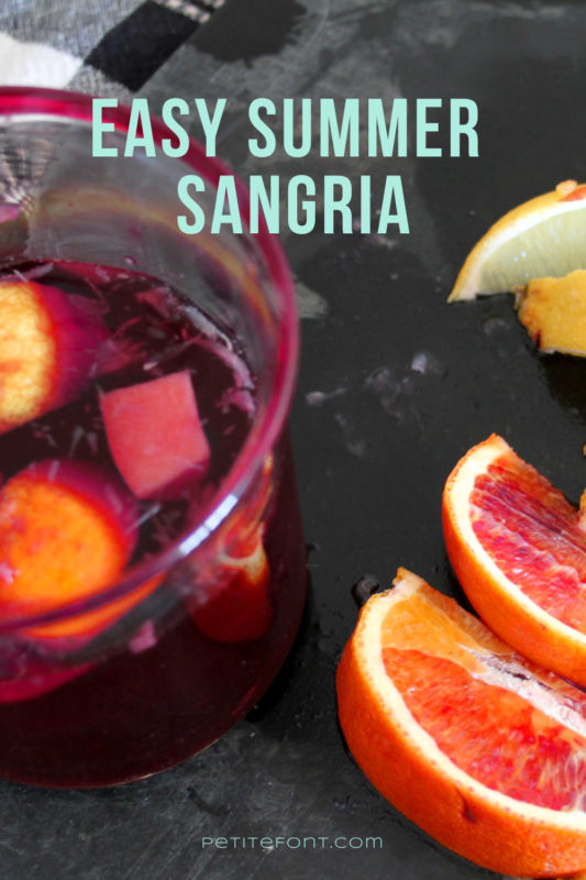 A zoomed in overhead view of a glass of sangria on a black wooden cutting board with blood orange and lemon wedges cut open on top of it. Text overlay reads "Easy Summer Sangria, PetiteFont.com"