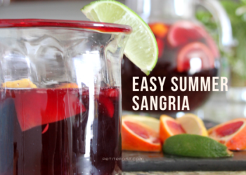 Glass on counter filled halfway with sangria, garnished with a lime wedge. Behind it is a bunch of cut up citrus, and a pitcher of the rest of the sangria. Text overlay reads Easy Summer Sangria, PetiteFont.com