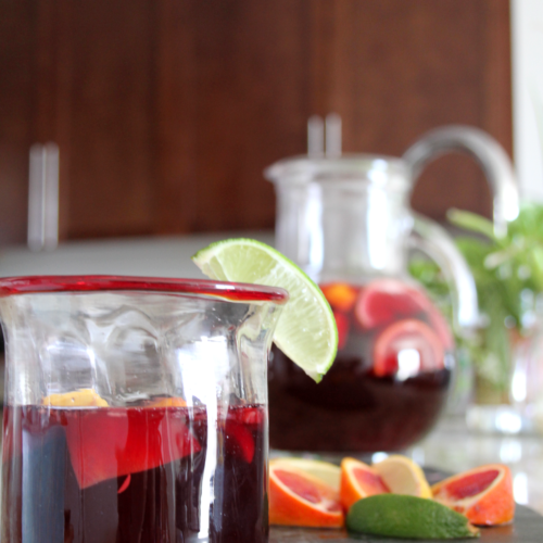 Glass on counter filled halfway with sangria, garnished with a lime wedge. Behind it is a bunch of cut up citrus, and a pitcher of the rest of the sangria