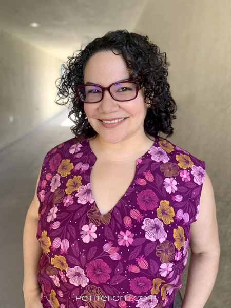 Latina woman with black curly hair and red glasses smiling at the camera in a floral open keyhole blouse