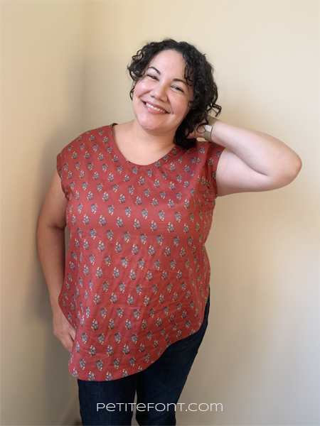 Curly haired Latina woman in a dark rust colored floral woven t-shirt with one hand up behind her head