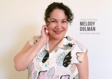 Paulette in front of a white wall, one hand up to her face, smiling. She is wearing a white button up camp shirt with a large scale kitschy pastel print of leaves and flowers. Text to the right of her reads Love Notions Melody Dolman Pattern Review.