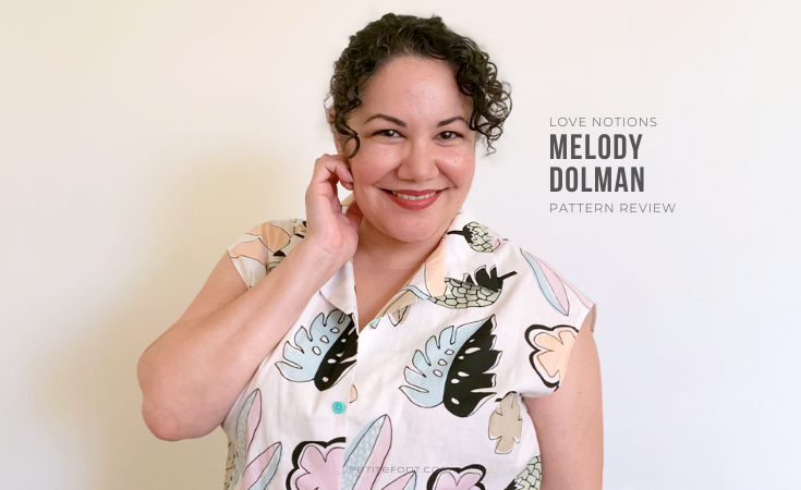 Paulette in front of a white wall, one hand up to her face, smiling. She is wearing a white button up camp shirt with a large scale kitschy pastel print of leaves and flowers. Text to the right of her reads Love Notions Melody Dolman Pattern Review.