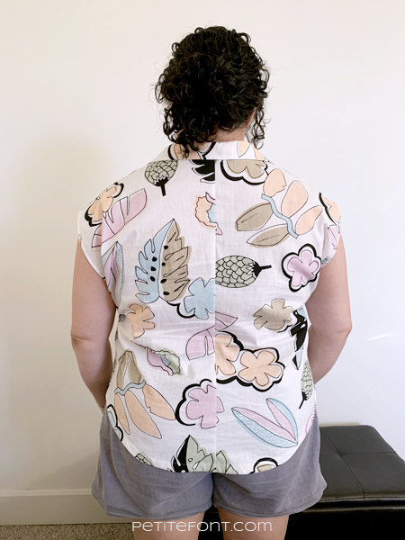 A back view of Paulette in front of a white wall and brown ottoman. She is wearing a white button up Melody Dolman with a large scale kitschy pastel print of leaves and flowers.
