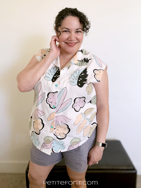 Paulette in front of a white wall and brown ottoman, one hand up to her face, smiling. She is wearing a white button up Melody Dolman with a large scale kitschy pastel print of leaves and flowers.
