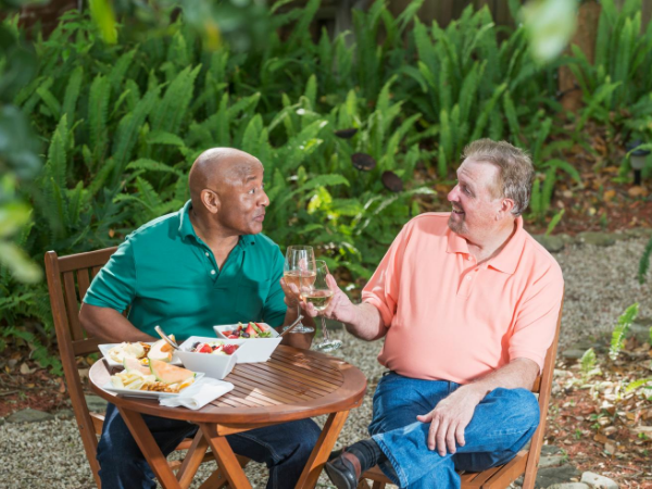 Two older men, one bald and black and one white with a goatee, sharing wine and food at a table in a garden