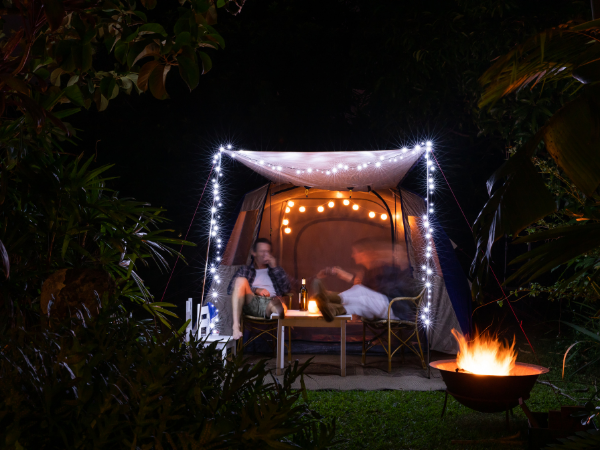 Night scene in back yard. A fire pit in the lower right corner with two people sitting in chairs under a lit canopy. A camping tent is behind them.