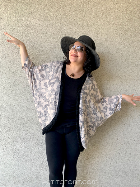 Paulette looking up to the left wearing a black hat and sunglasses with her arms out like a bird. She is in a black shirt and pants with a peach paisley version of Simplicity 1108 over the top.