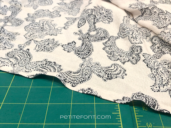 Zoomed in view of a peach fabric with a black paisley print on top of a green ruled cutting mat. It highlights the ripples along the stretched out edge of Simplicity 1108.
