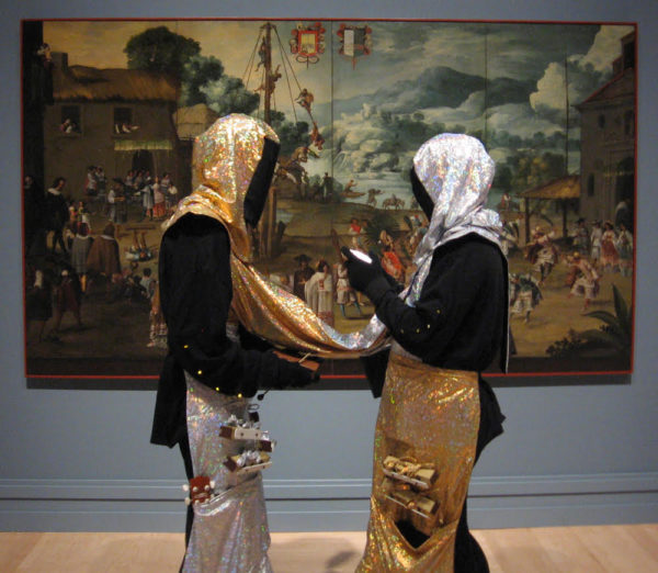 Two people standing in front of a large colorful fine art painting. They are facing each other and completely covered in black with gold and silver headcoverings, aprons, and a tube connecting them.