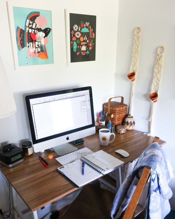 Corner desk with an Apple computer, open notebook, coffee mug and other desk tools on top. There are pretty prints on the wall above and macrame wall hangings to the right.