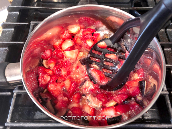Saucepan on the stove with macerated strawberries and figs being mashed by a potato masher