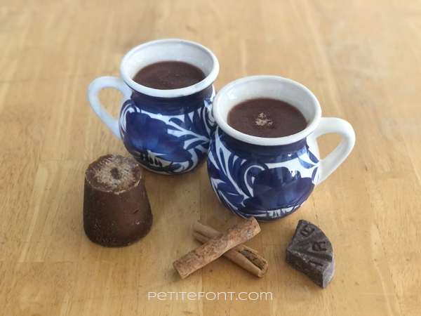 2 cups of champurrado in white mugs with a blue design, sitting on top of a wooden table. In front of the mugs are a used cone of piloncillo, 2 cinnamon sticks, and a wedge of Mexican chocolate