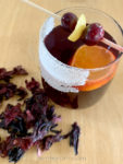 A glass of non-alcoholic mulled wine garnished with cranberries and lemon zest, dried hibiscus flowers scattered about